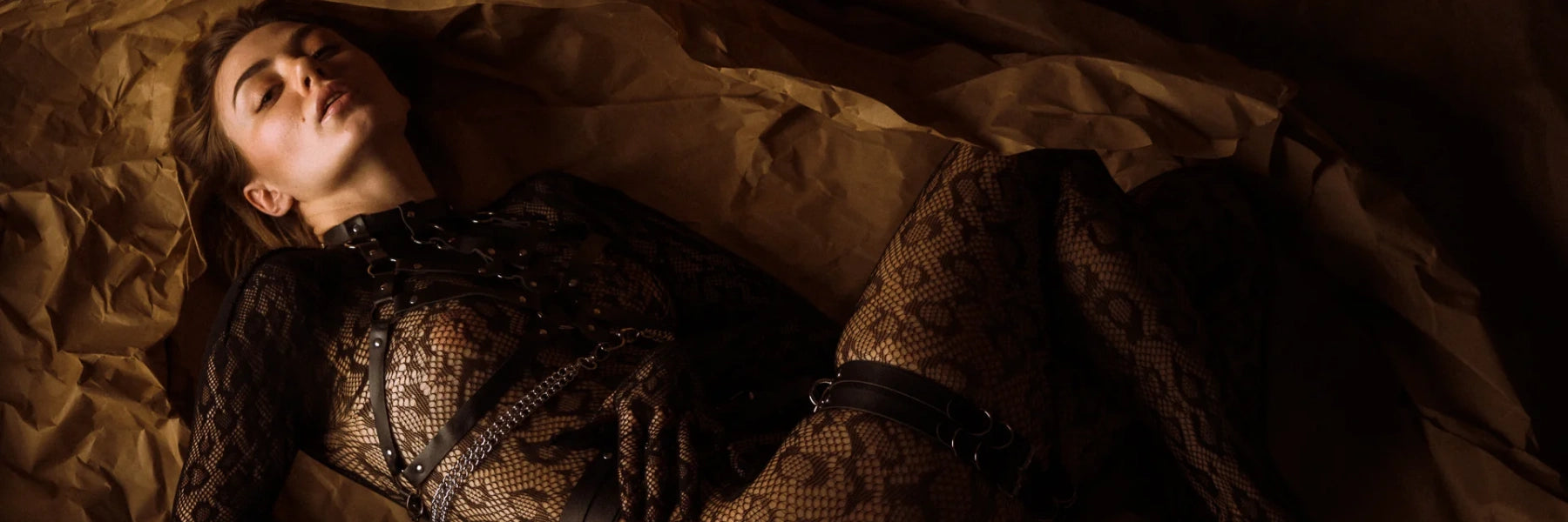 A seductive woman wearing a black lace bodysuit with leather chain harness, lying on textured brown paper under soft moody lighting — showcasing Loviy’s luxury fetish lingerie collection.